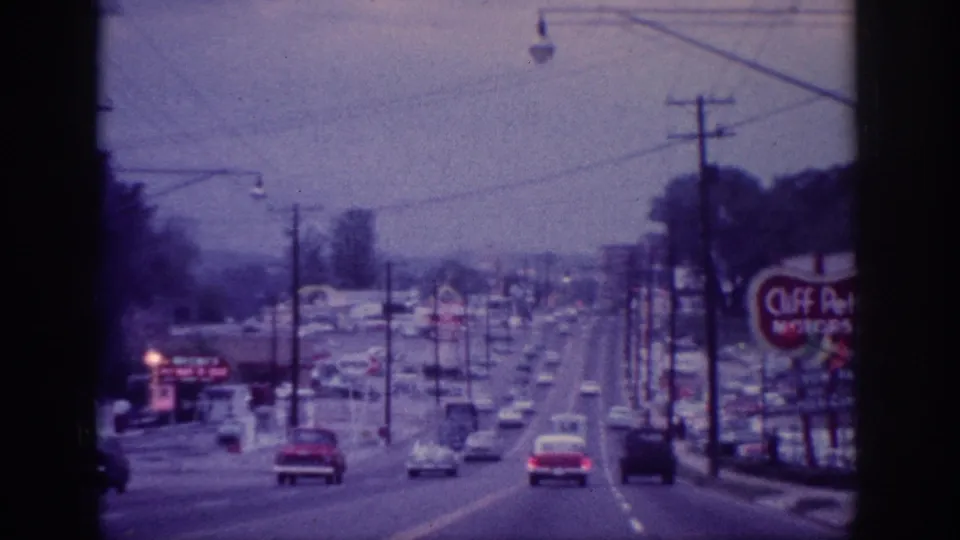Forward-facing 8mm view of Memphis boulevard traffic with several cars, overhead utility lines, streetlights, storefront signage, and a prominent roadside motel sign.
