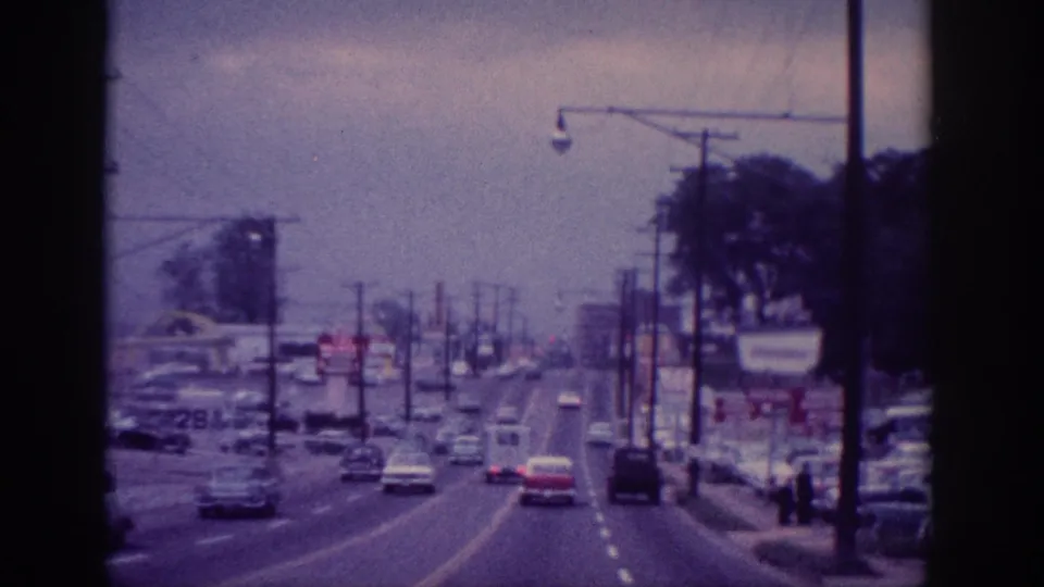 1963 Memphis driving view showing classic cars on a multi-lane road, roadside commercial signs, utility poles, and small distant pedestrians near the curb.