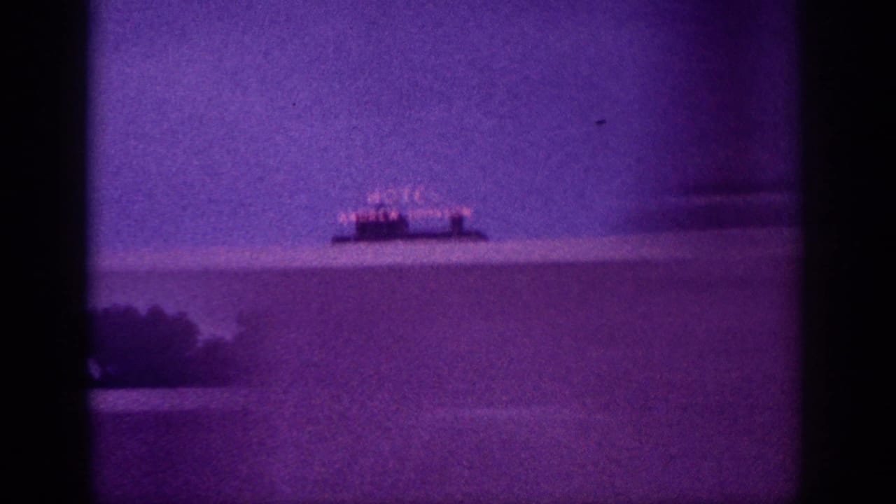Wide shot of broad water expanse with a small illuminated sign visible on the distant shore, heavy purple cast and film grain.