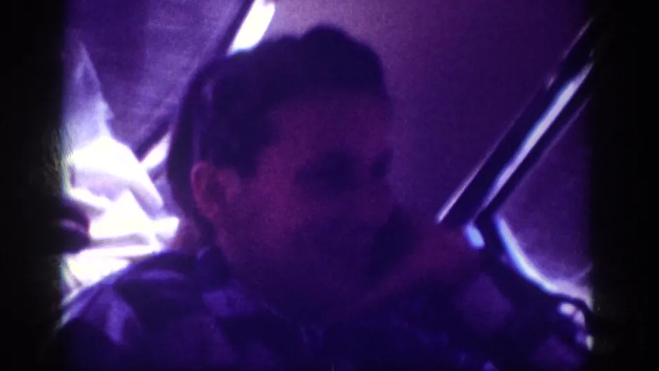 Young man in 8mm home movie turning in car backseat circa 1963, with strongly overexposed window light and car door frame visible behind him.