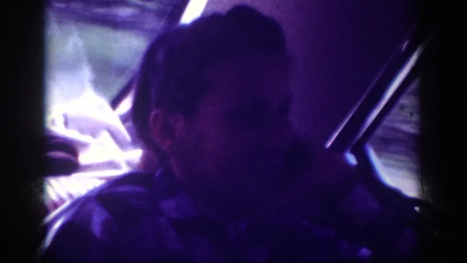 Young man in 8mm home movie circa 1963 seated in car backseat, looking toward window as faint exterior light filters through the glass.