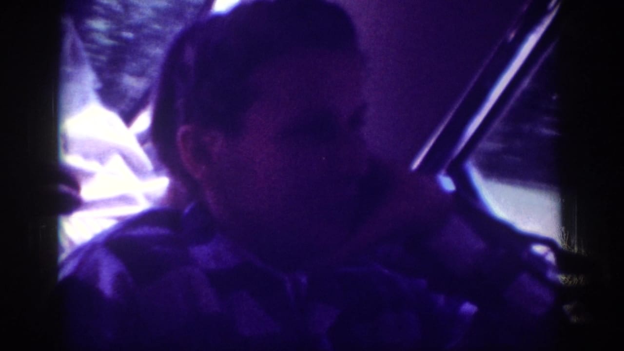 Adult man seen from rear-passenger angle inside a moving car, heavy blue-violet color cast from aged 8mm film.