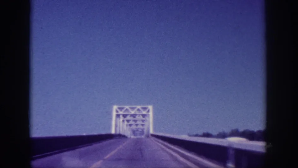 Forward approach toward a white steel truss bridge along a two-lane road, ca. 1963, Memphis Tennessee area; shot on 8mm film.