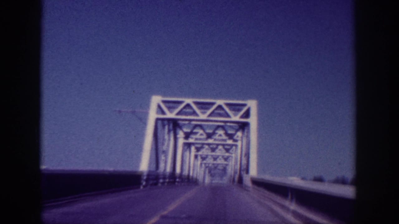 1963 Memphis vehicle POV approaching a steel truss bridge, with the first white truss portal ahead under a blue sky.