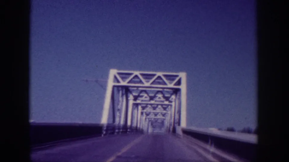 1963 Memphis vehicle POV approaching a steel truss bridge, with the first white truss portal ahead under a blue sky.