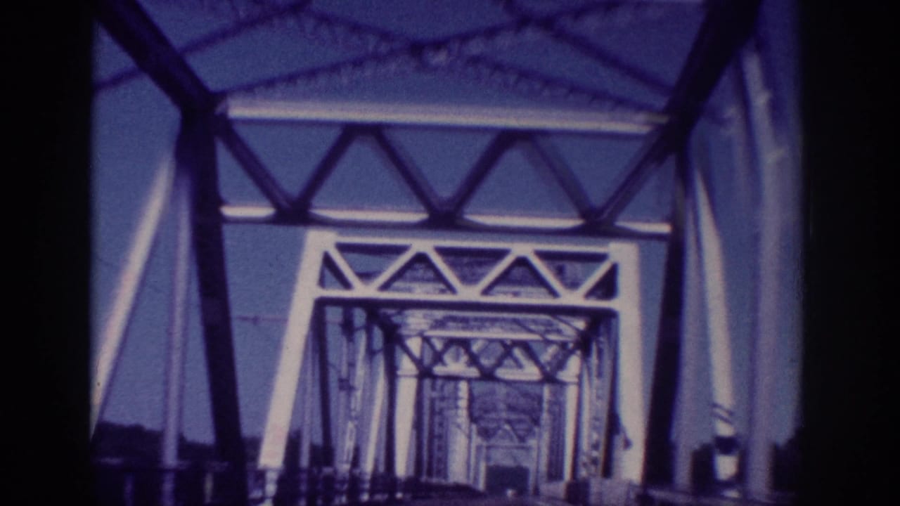 1963 Memphis home-movie frame looking forward from a car into a steel truss bridge, with dark film edges and blue-purple color shift.
