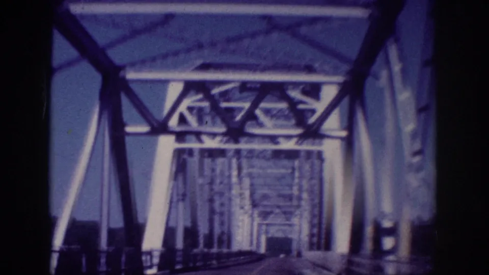 Forward-looking 8mm frame from inside a moving car on a Memphis steel truss bridge, showing the bridge structure narrowing into the distance.