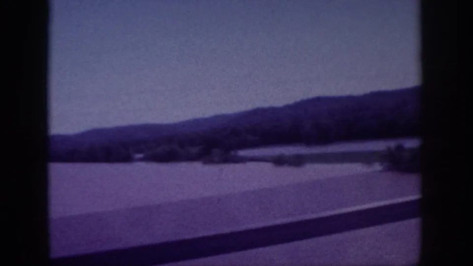 1963 Memphis travel frame looking from the bridge toward a broad body of water, low wooded hills, shoreline, and roadside railing.