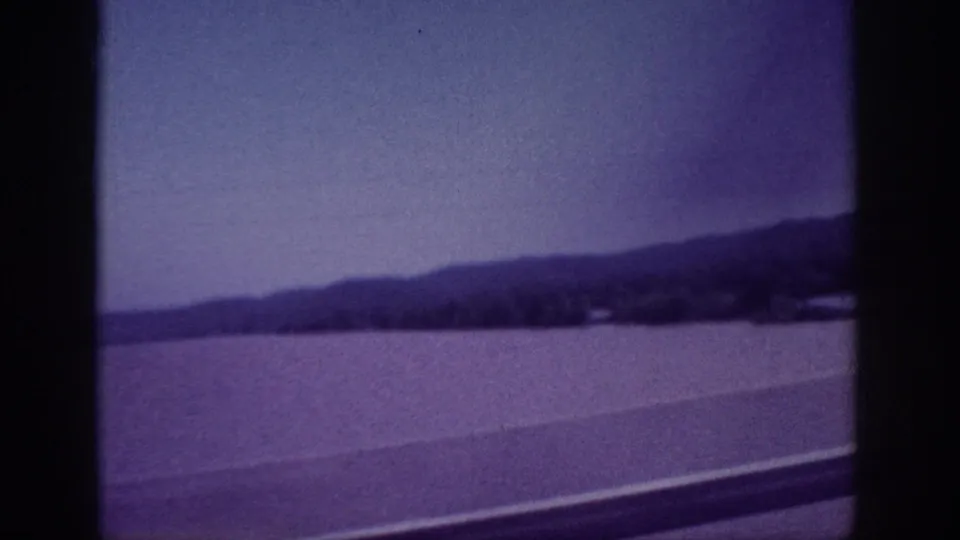 Soft 8mm frame from a moving car on a Memphis bridge, showing water, distant wooded hills, shoreline, and the lower bridge railing.