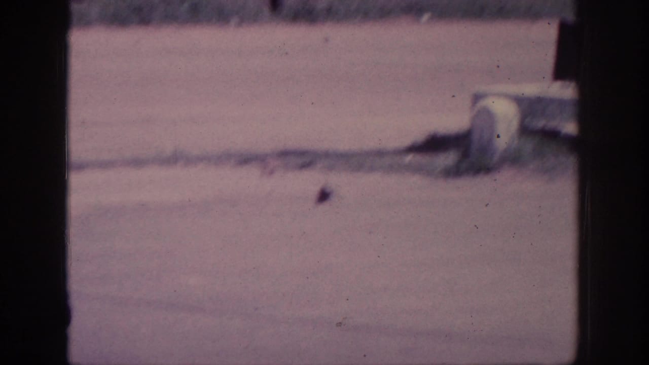 Wide dusty road in 1963 8mm home-movie footage; a small dark object rests on the sunlit pavement near a white roadside cylinder.