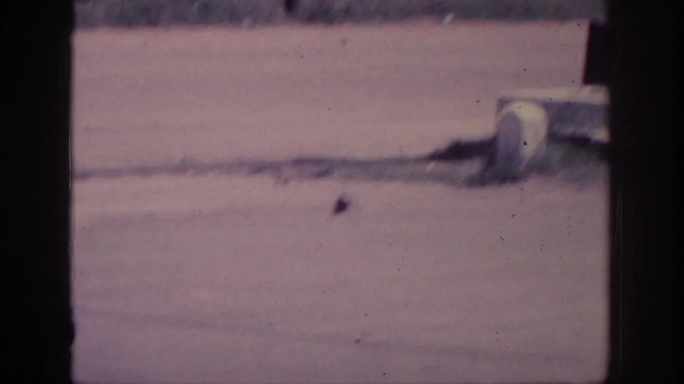 Wide dusty road in 1963 8mm home-movie footage; a small dark object rests on the sunlit pavement near a white roadside cylinder.