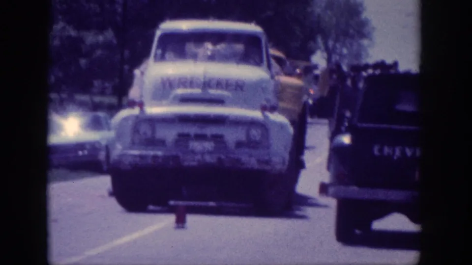 1960s white commercial truck and dark Chevrolet pickup on a sunlit US highway, shot on 8mm; partial lettering on truck cab; tree line behind both vehicles.