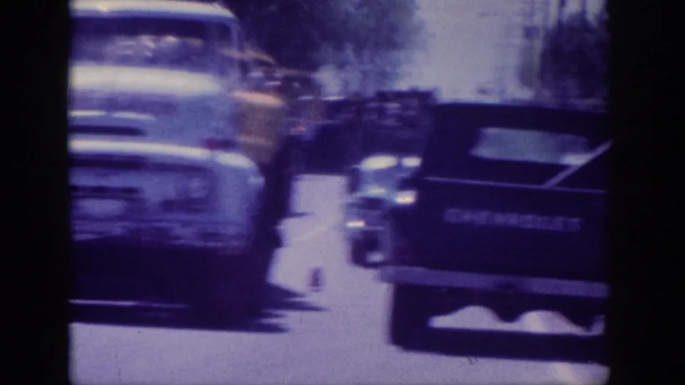 Extreme motion blur from handheld 8mm filming as period trucks pass; white cab and dark Chevrolet pickup remain partially identifiable shapes within the blur.