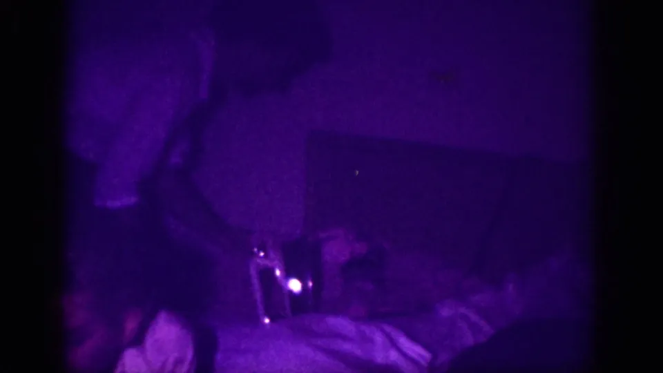 8mm home movie frame, ca. 1963, Memphis, Tennessee; purple-cast low-light interior with a partial figure above a flat surface and a small bright point, possibly an iron indicator lamp.