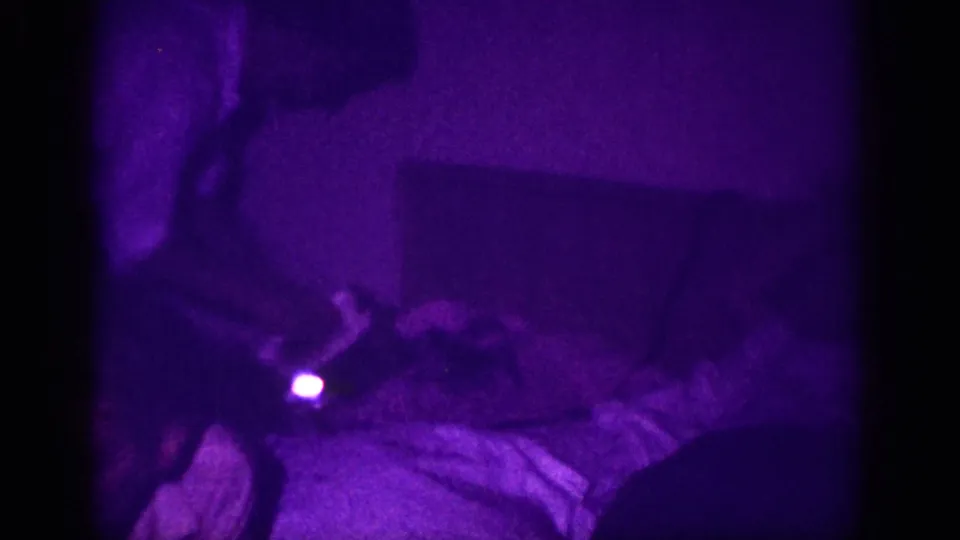 8mm home movie frame, ca. 1963, Memphis; heavily purple-cast interior with what may be garments on a bed or table, a small bright indicator light near the lower center, and a figure partially visible at top.
