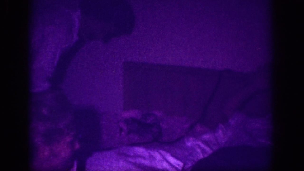 8mm home movie frame circa 1963 showing a figure reclining on a bed in a bedroom suffused with intense purple color cast from aged film dye degradation