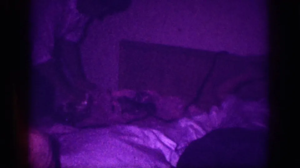 8mm home movie frame circa 1963 showing two figures in a bedroom with rumpled bedding, dominated by a purple-violet color cast from aged 8mm film stock