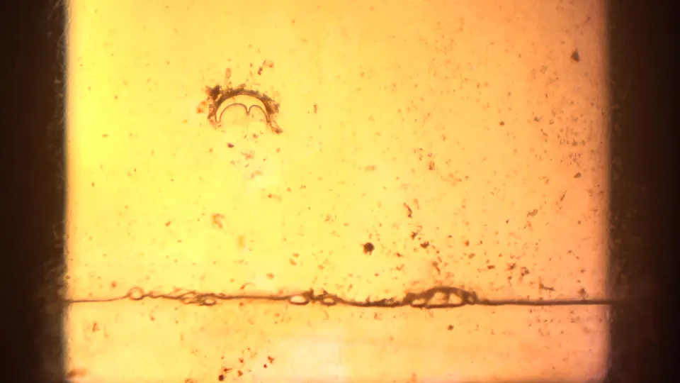 End section of 8mm film stock showing amber-yellow fogging with visible dust, debris particles, a splice or tear mark, and physical film deterioration
