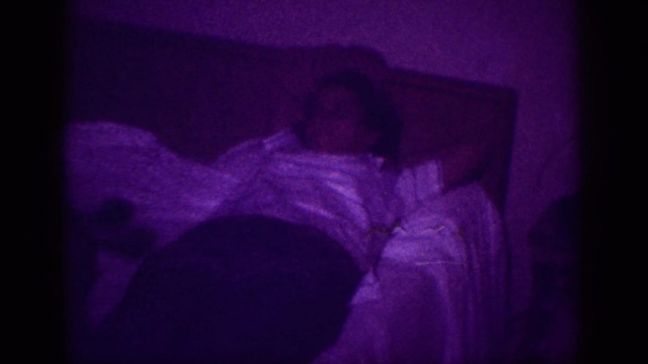 Person at rest in bed, white blanket visible, dark headboard behind; heavy purple color shift from 8mm film processing anomaly, Memphis 1963.