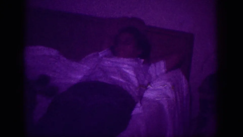 Person at rest in bed, white blanket visible, dark headboard behind; heavy purple color shift from 8mm film processing anomaly, Memphis 1963.