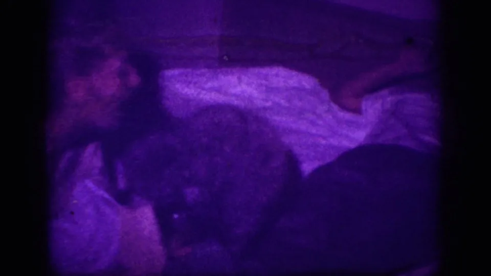 Person lying in bed with white blanket; dark form at right consistent with a dog; strong purple color cast from 8mm film anomaly, circa 1963.