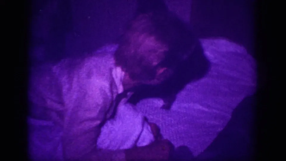 Person and dark dog lying close together on bed with white blanket; purple processing anomaly and coarse grain from 8mm home movie, circa 1963.