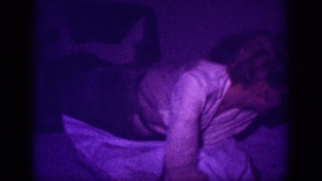 Woman in white blouse leaning forward over a bed with white bedding, dim interior room, heavy purple color cast.