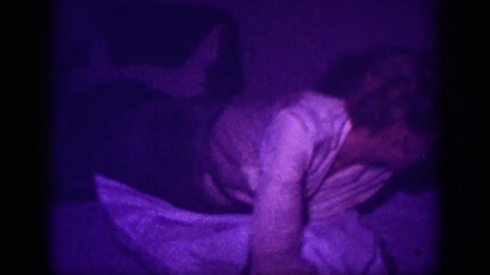 8mm home movie frame circa 1963, Memphis, Tennessee: woman in white blouse bent forward over a bed in a dim interior saturated with purple dye shift.