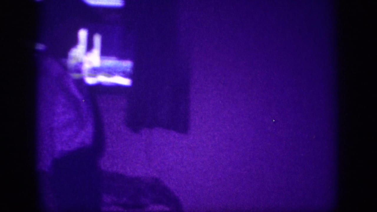 8mm home movie frame circa 1963: interior room suffused with purple dye shift, a figure's raised arm visible with small objects on a shelf or dresser in the upper background.