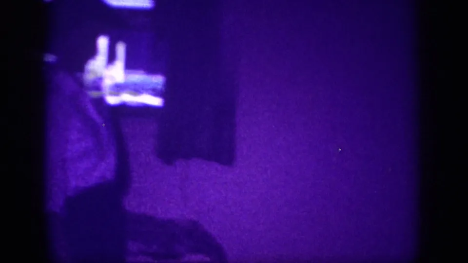 8mm home movie frame circa 1963: interior room suffused with purple dye shift, a figure's raised arm visible with small objects on a shelf or dresser in the upper background.