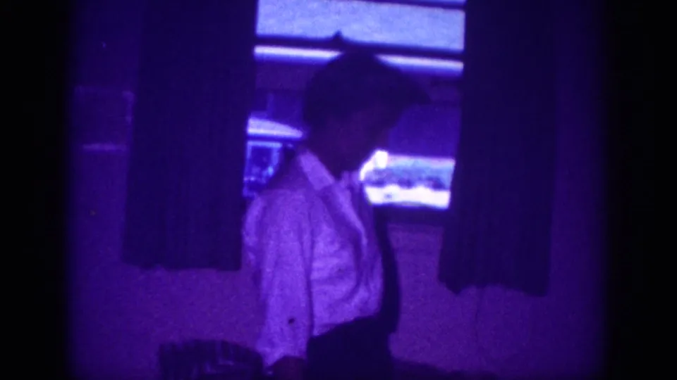 8mm home movie frame circa 1963, Memphis, Tennessee: woman in white blouse standing before horizontal window blinds flanked by dark curtains, room saturated in purple.