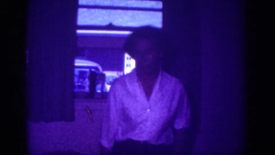 8mm home movie frame circa 1963, Memphis, Tennessee: woman in open-collar white blouse near window blinds, turning toward camera with domestic furnishings behind her.