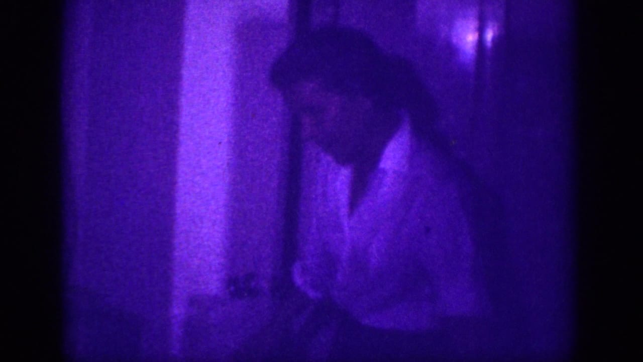 Man in white shirt pausing mid-movement in a purple-lit interior, wall corner visible behind him.