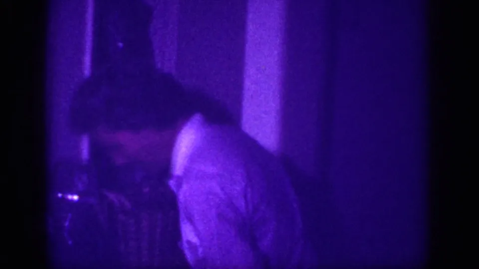 Two adults visible in a purple-tinted domestic interior, foreground figure in a checked shirt; 8mm home movie footage attributed to Memphis, Tennessee, circa 1963.