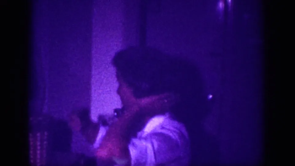Close view of two adults in a purple-lit domestic interior, one appearing to reach toward a table; 8mm home movie footage attributed to Memphis, Tennessee, circa 1963.