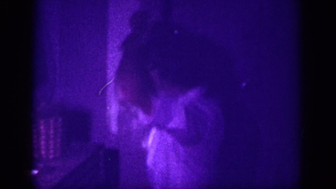 Woman in light blouse stands in a darkened interior room, entire frame dominated by heavy purple-violet color cast from aged 8mm film.