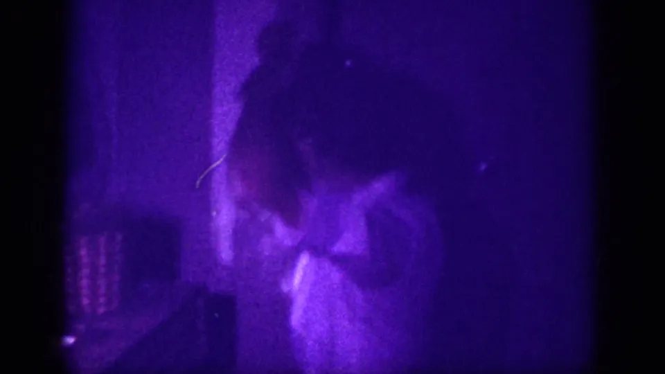 Woman in a light-colored blouse in a dimly lit domestic interior, captured on 8mm color film with advanced purple dye shift, Memphis Tennessee area, circa 1963.