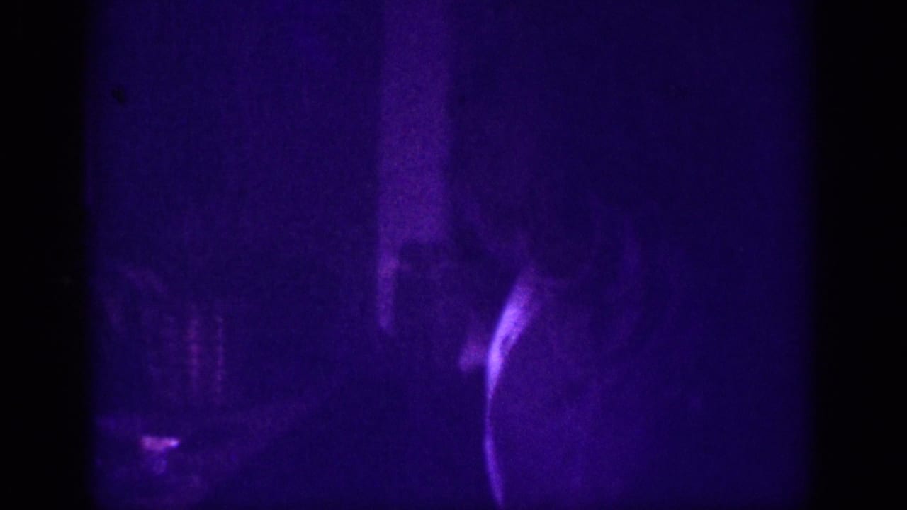 Upper body of woman barely visible against dark background, purple-tinted 8mm frame with indistinct objects faintly visible at right.