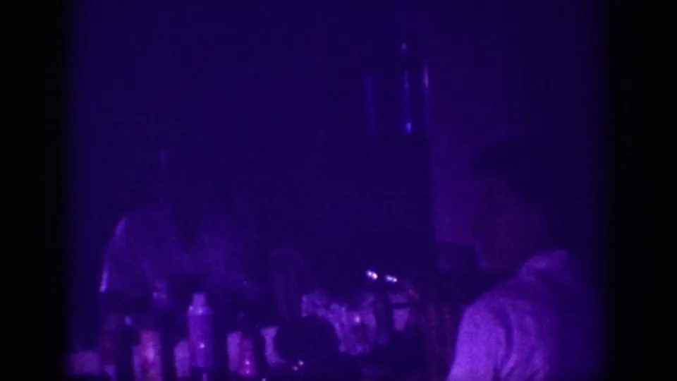 8mm home movie frame from 1963 Memphis: a woman's silhouette near a counter of bottles, everything cast in deep purple UV light with heavy film grain.