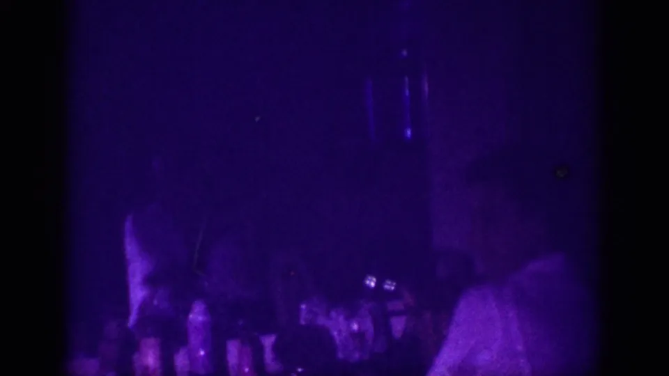 8mm home movie frame from 1963 Memphis: a woman leans toward a counter of bottles in a room illuminated entirely by deep purple UV blacklight.