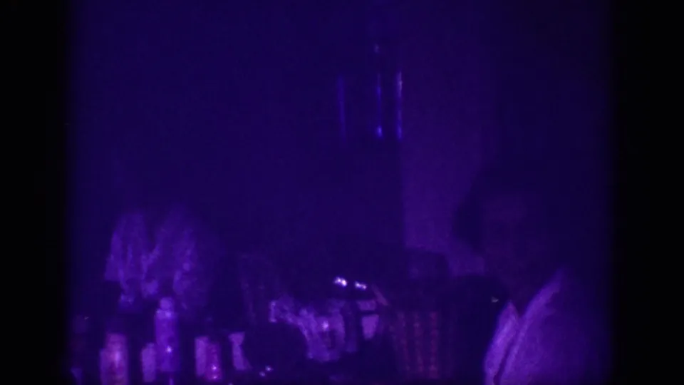 8mm home movie frame from 1963 Memphis: woman's silhouette near hair care products on a counter, lit by purple UV blacklight with pronounced film grain.
