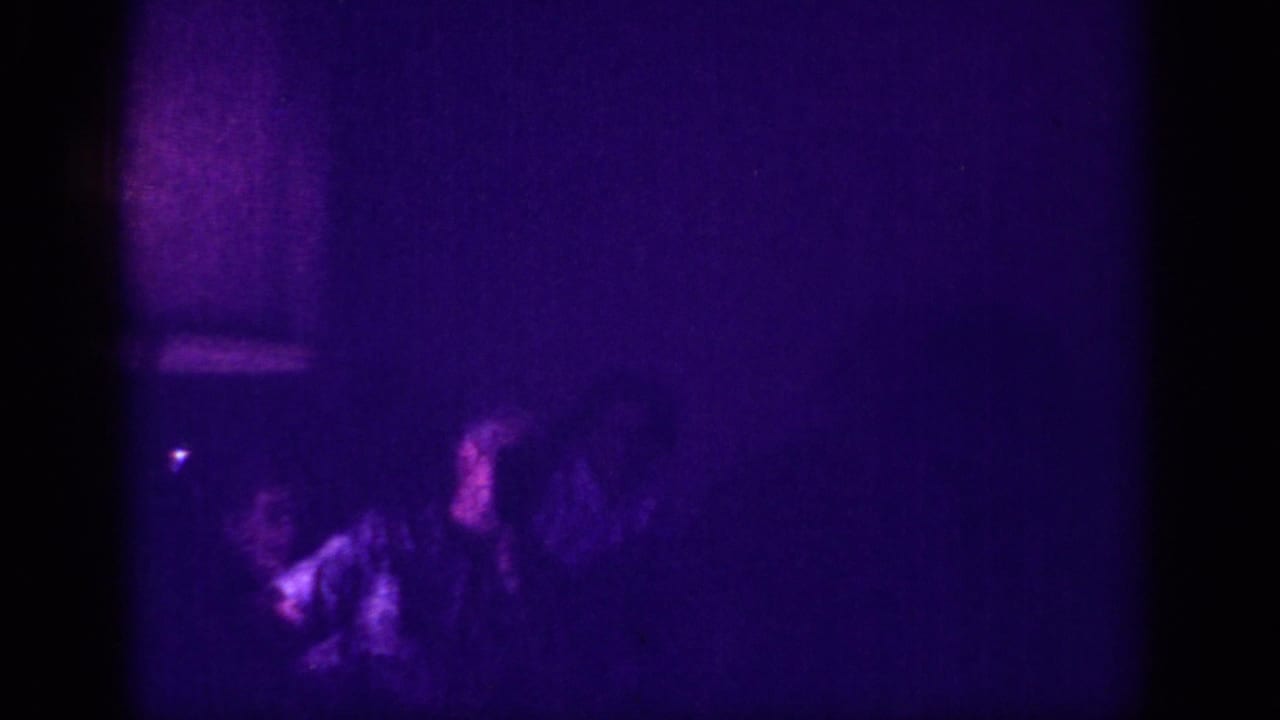 Two or three silhouetted figures in a dim purple-lit interior; one in a light-colored garment, a small point of light visible.