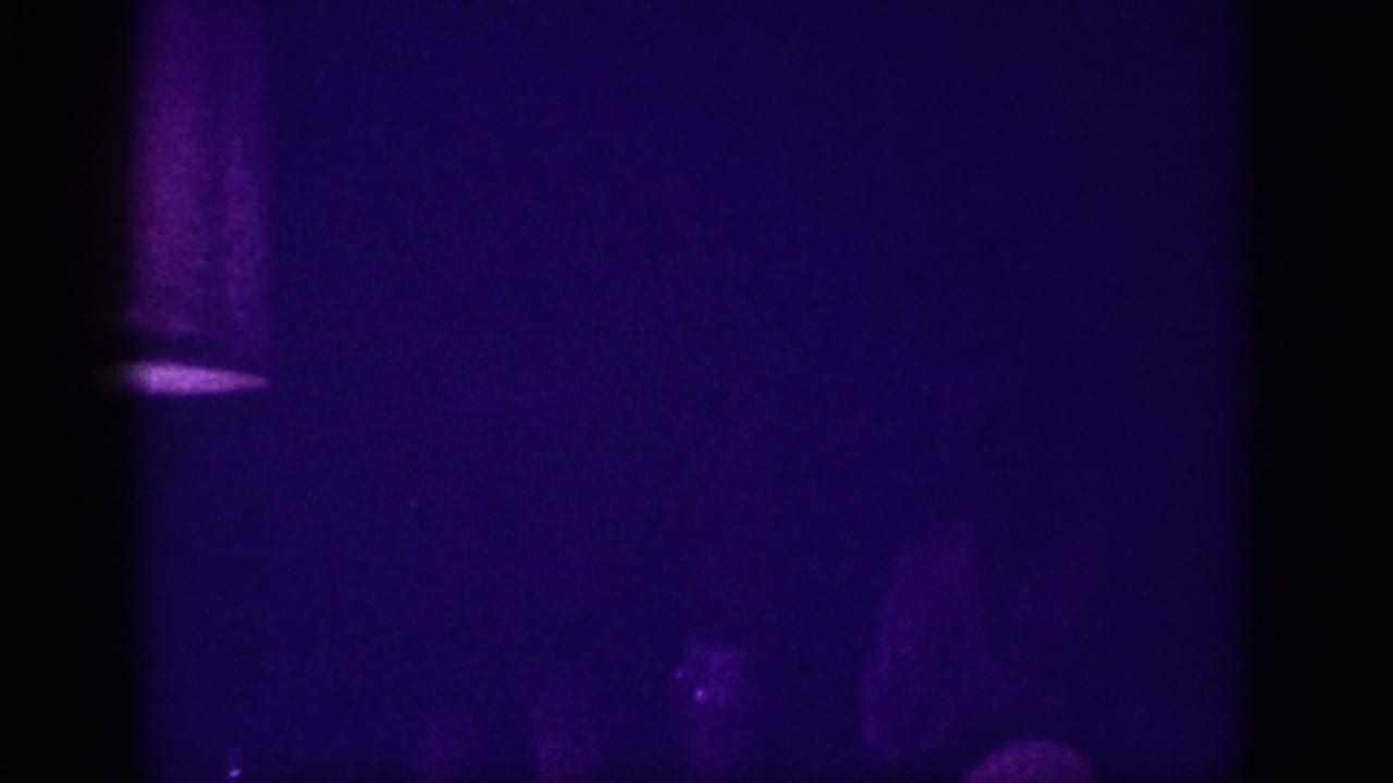 Extremely dark, deep purple-tinted 8mm film frame showing a barely visible interior scene with no discernible detail.