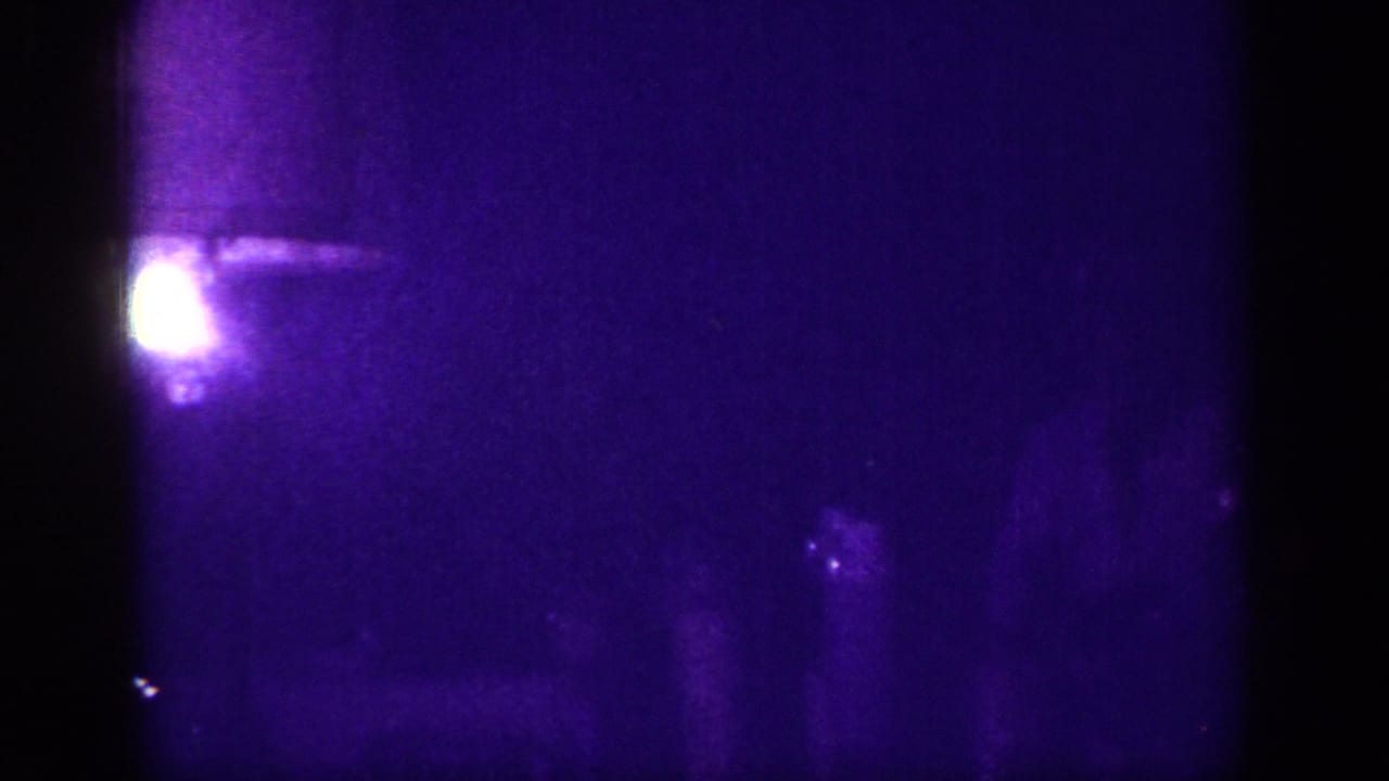 Severely underexposed 8mm interior with a glowing light source — possibly a lamp or fixture — and a barely visible standing figure against a deep purple-blue background, circa 1963.