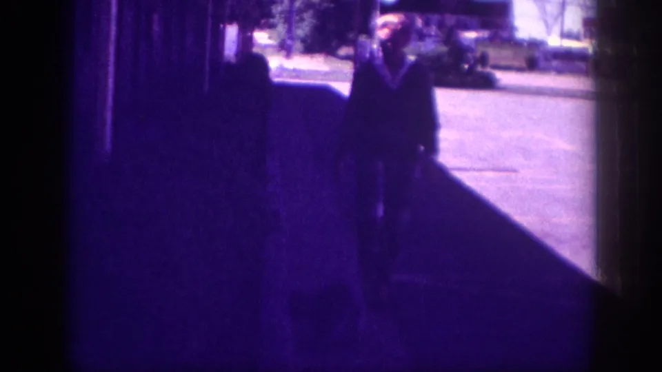 A dark-clothed figure on a residential sidewalk in daytime; trees, a street, and faint unreadable background signage visible in what may be Memphis, Tennessee, circa 1963.
