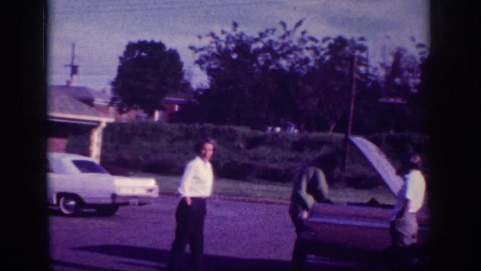 Two women in white shirts and dark pants beside a 1960s American sedan with open trunk in a suburban driveway, Memphis Tennessee, 1963