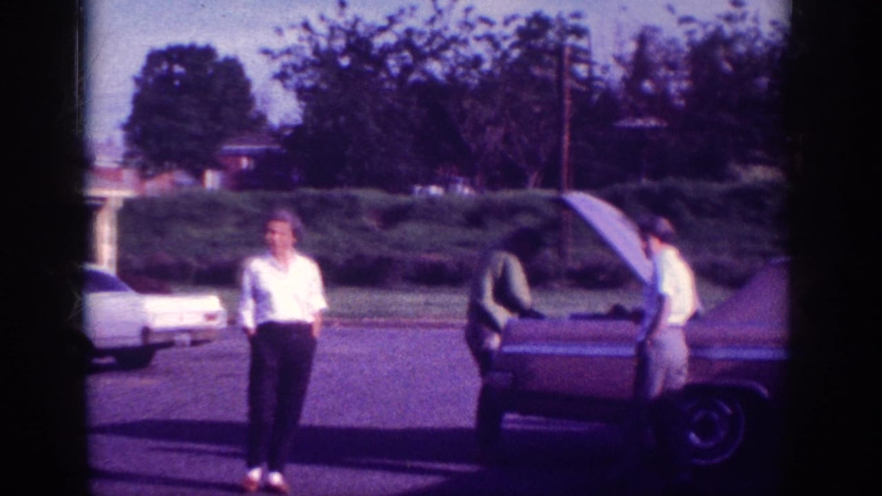 Woman in white shirt standing near rear of large dark American sedan with trunk raised, suburban street, 8mm film