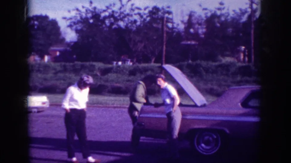 Two women examining the trunk area of a large 1960s American sedan in a suburban driveway, Memphis Tennessee, circa 1963, on aged 8mm color film