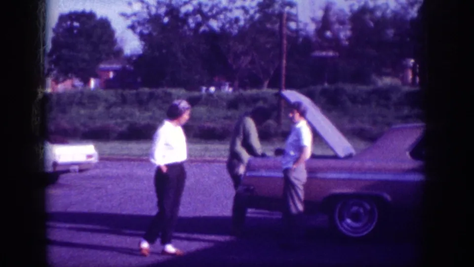 Two women in period attire standing beside the rear of a 1960s American sedan on a suburban street, Memphis Tennessee, 1963