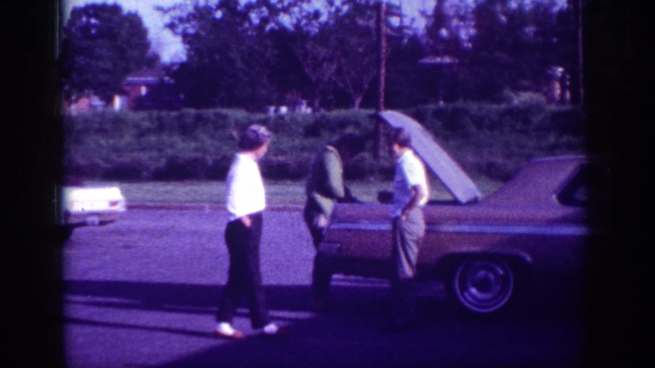 Three men standing around a large 1960s American sedan with the hood raised on a suburban street.